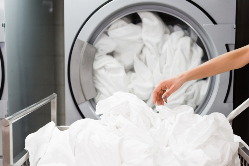 A person’s hand reaches toward a large industrial washing machine filled with freshly laundered white linens, with a cart of clean laundry in the foreground.
