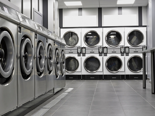 A modern laundromat features rows of industrial front-loading washers and dryers in a sleek, clean space with gray tile flooring and bright overhead lighting.

