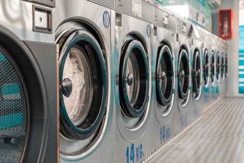 A row of large-capacity, stainless steel washing machines lines the wall of a modern laundromat, with one machine actively washing a load of laundry.