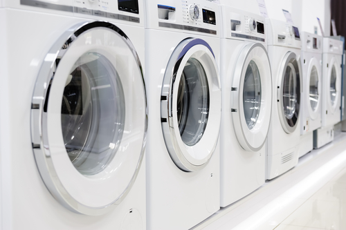 A row of modern front-loading washing machines is neatly lined up in a bright, clean appliance showroom.
